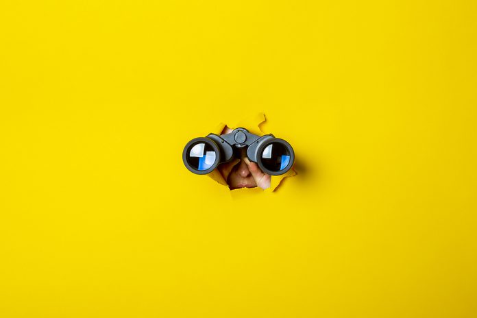 Female Hand Holds Black Binoculars On A Yellow Background. Journ